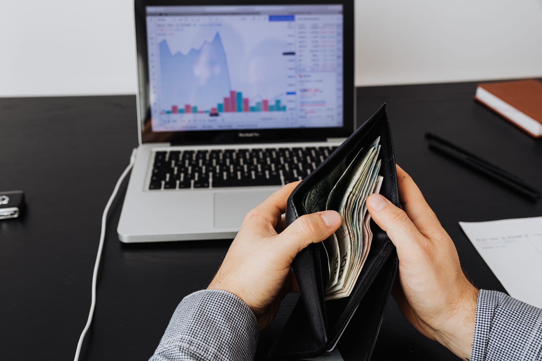 Man holding a leather wallet with bills in front of a laptop displaying financial graphs.